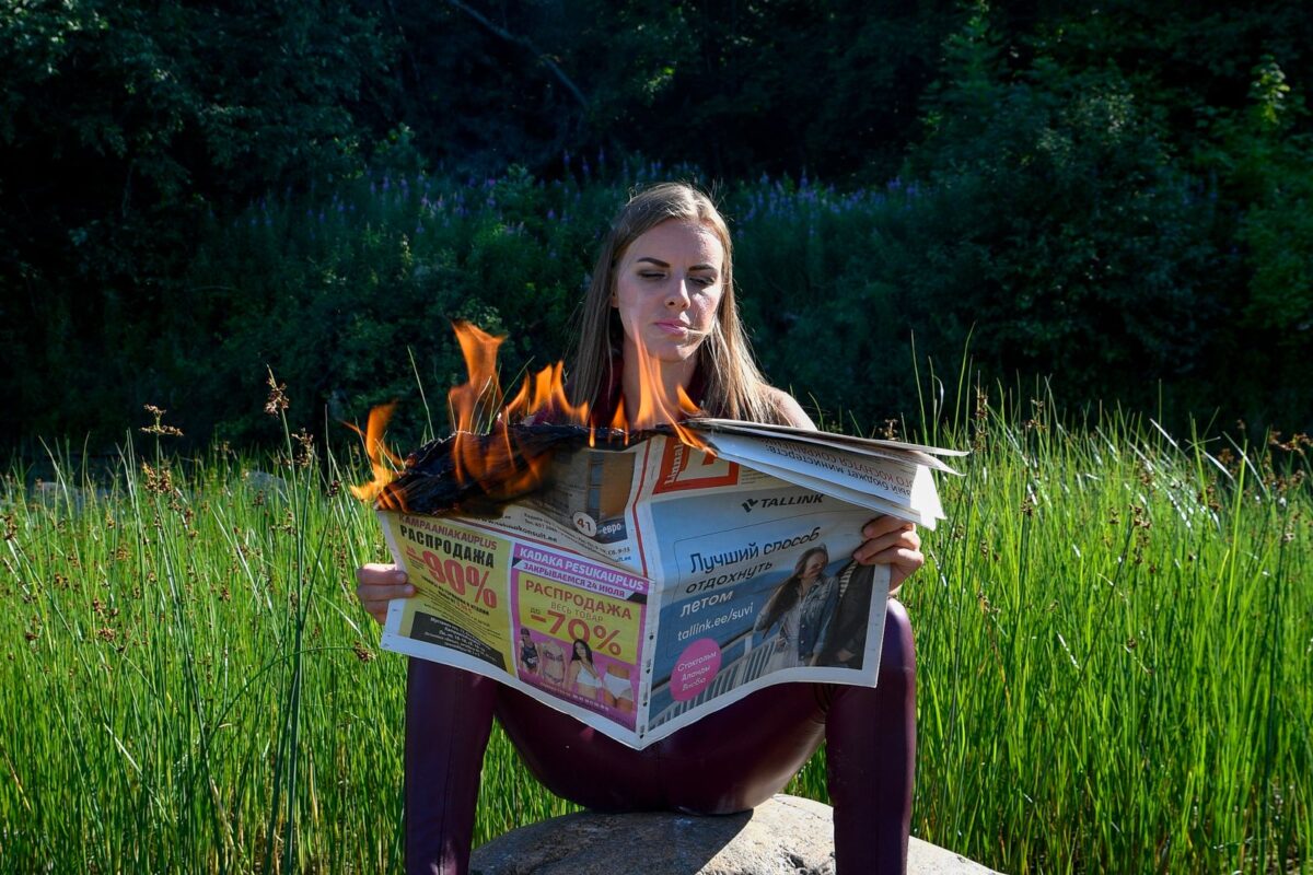 A young woman calmly reads a flaming newspaper while sitting in a green field.